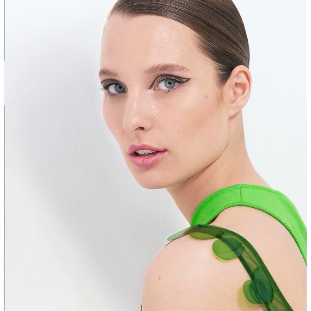 Close-up portrait of a fair-skinned woman with blue eyes, wearing green dress, and dark eyeliner against a white background.