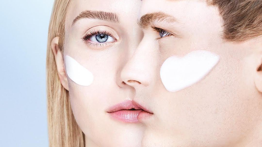 Close-up of a woman and man's faces with white cream on their cheeks in a studio setting, showcasing skincare and beauty.