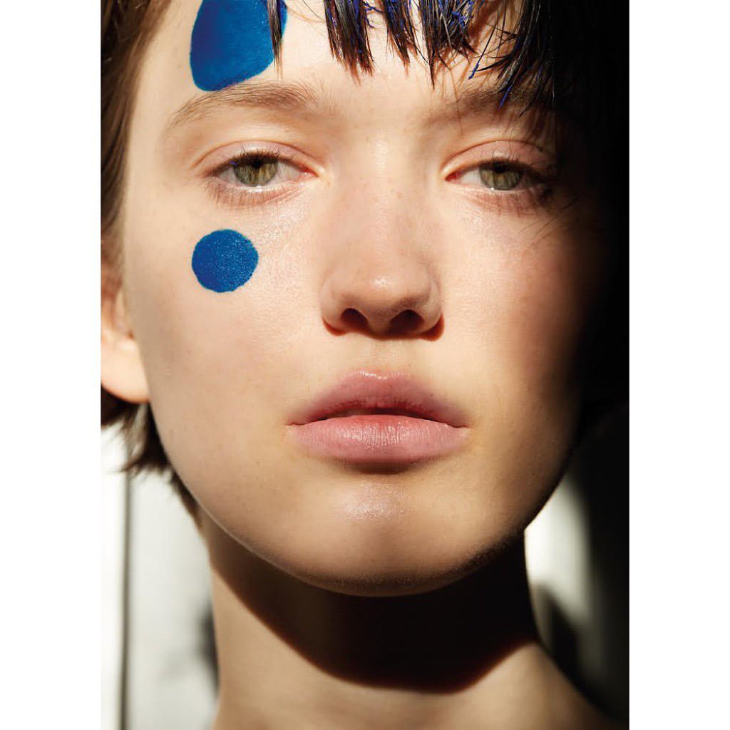 Close-up portrait of a fair-skinned young woman with short hair and blue paint on her face, gazing directly at the viewer.