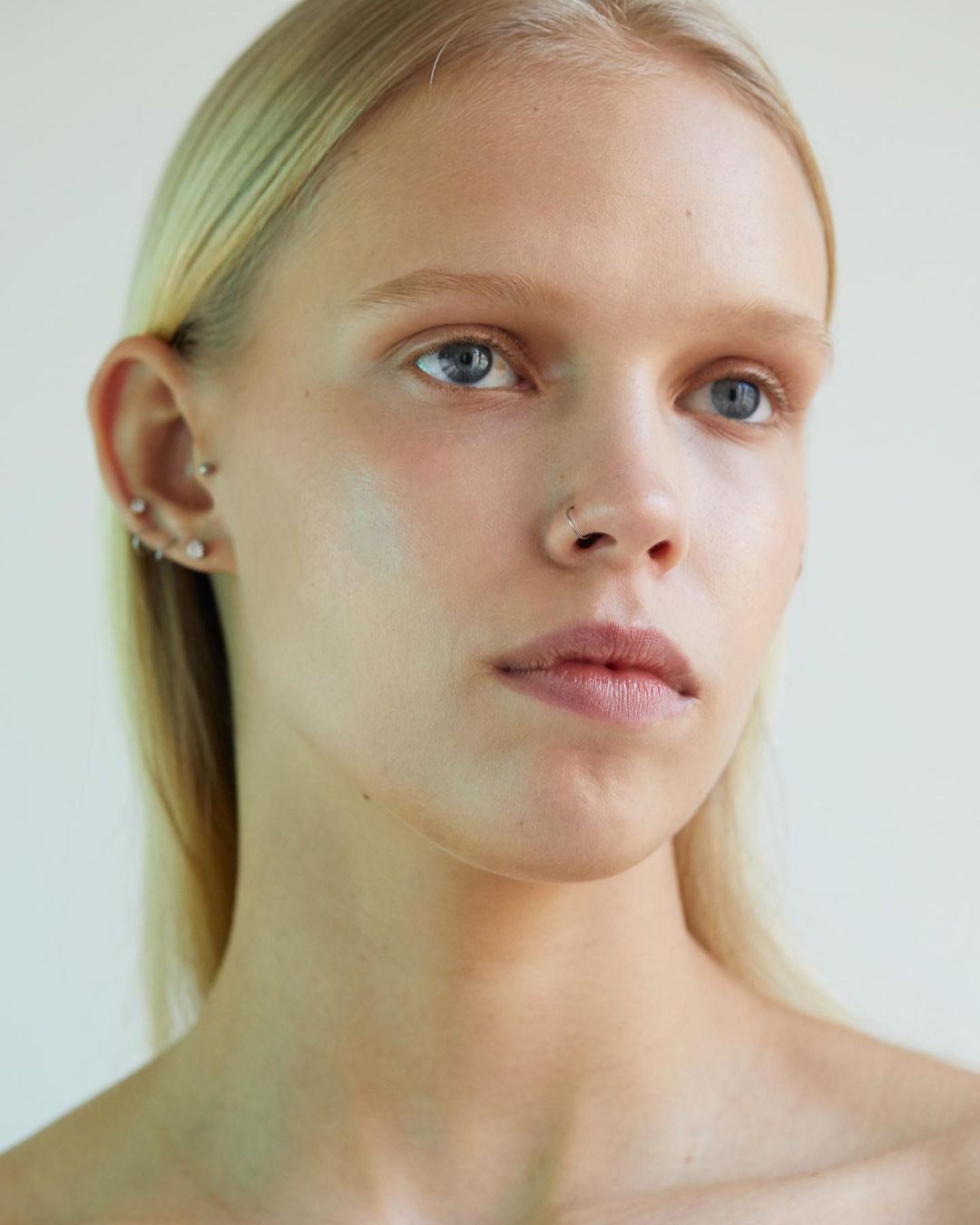Close up portrait of a young woman with blonde hair, light eyes, and facial piercings against a plain neutral background.