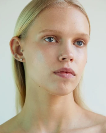 Close up portrait of a young woman with blonde hair, light eyes, and facial piercings against a plain neutral background.