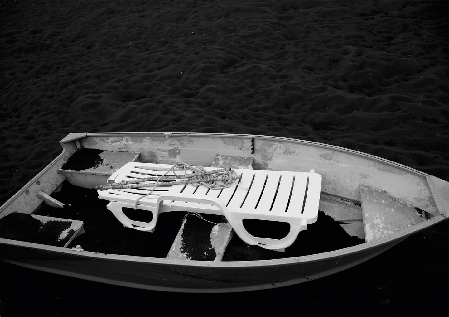 Monochromatic view of an empty boat containing a white plastic lawn chair resting on a dark surface within, floating on the water.