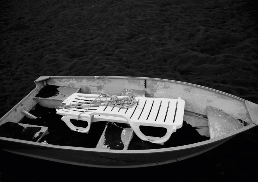 Monochromatic view of an empty boat containing a white plastic lawn chair resting on a dark surface within, floating on the water.