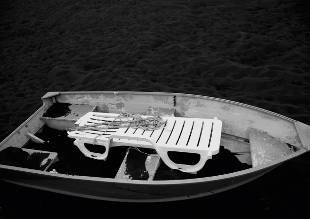 Monochromatic view of an empty boat containing a white plastic lawn chair resting on a dark surface within, floating on the water.