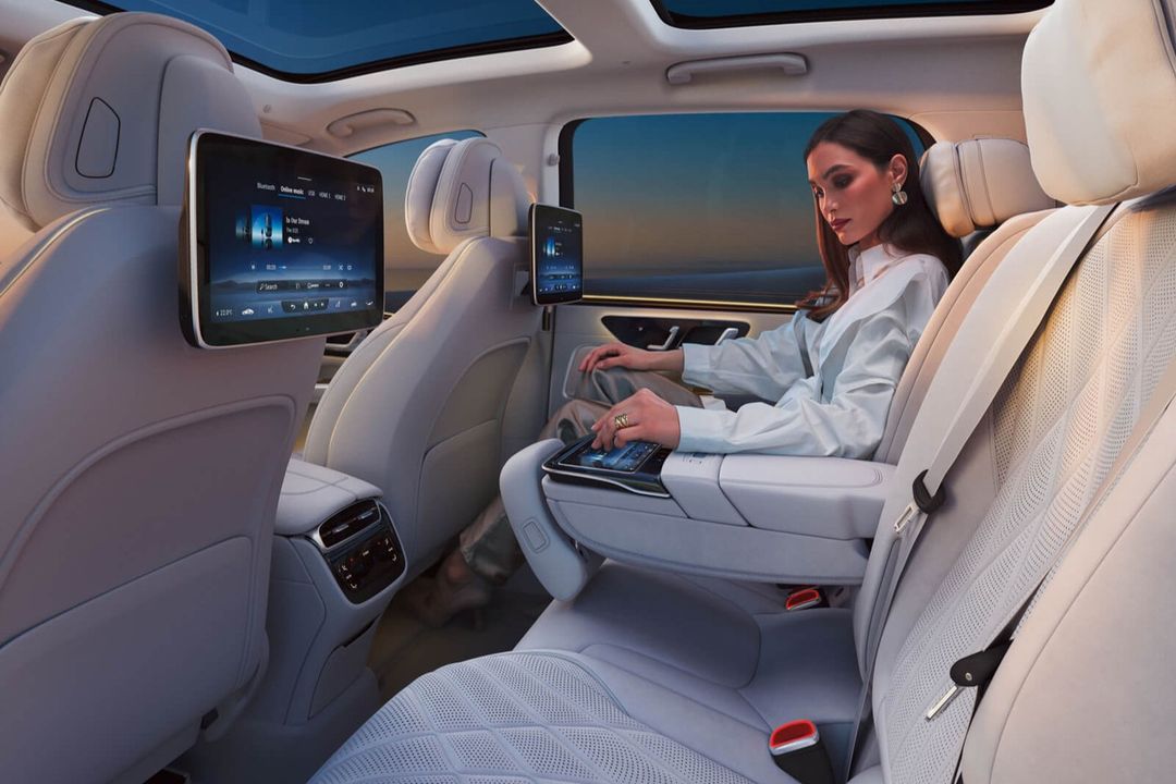 Adult female sits in the back of a luxury car with screens on the seatbacks. She is wearing a white blouse and using a tablet.