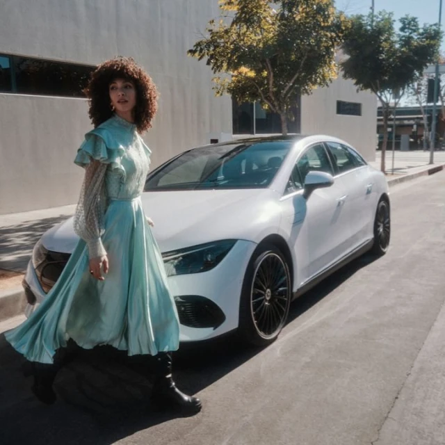 A woman in a long blue dress walks next to a white Mercedes-Benz car on a city street with buildings and trees in the background.