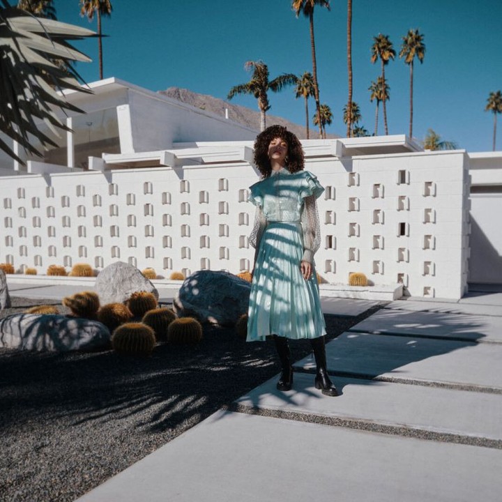 A woman with curly hair in a teal dress stands on a concrete path, with a white building, palm trees, and mountains in the background.