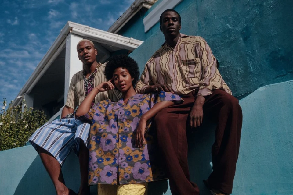 Three stylish African models pose outdoors wearing patterned shirts and brown trousers against a blue wall and blue sky backdrop.