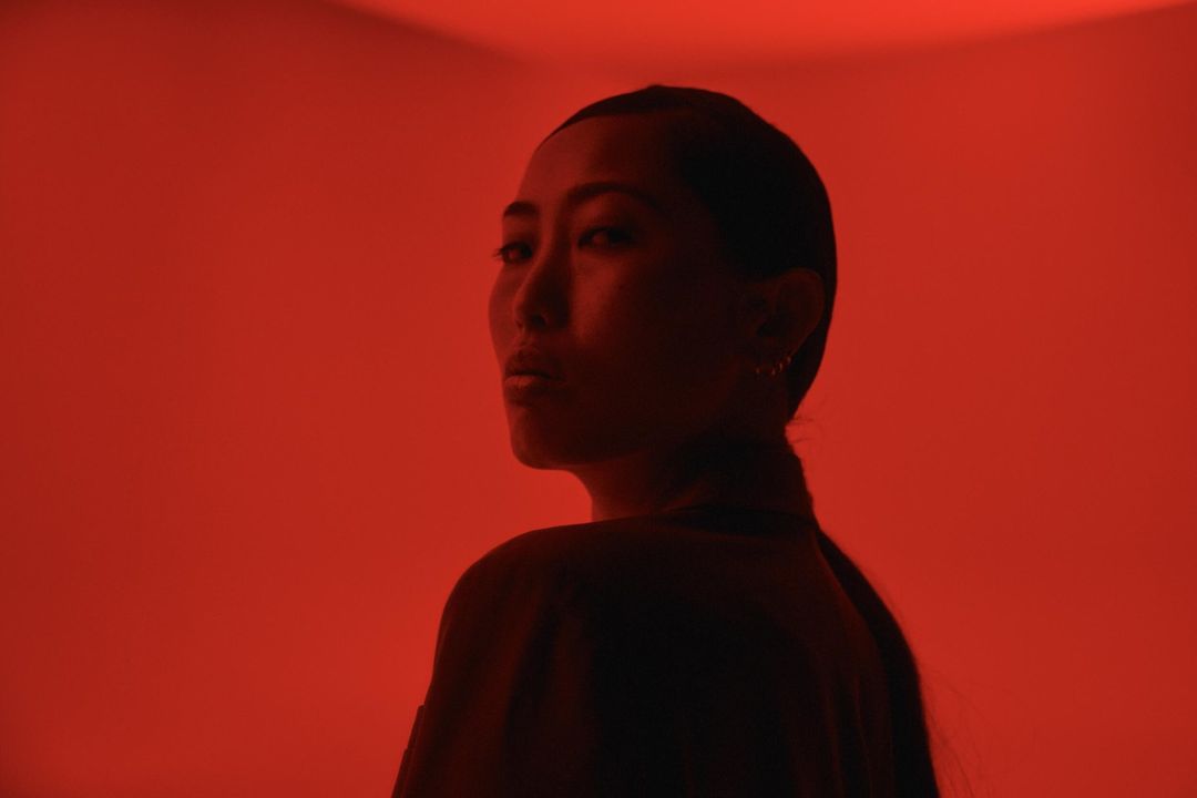 Portrait of an adult woman with dark hair looking over her shoulder in a photo studio bathed in red lighting, casting dark shadows.