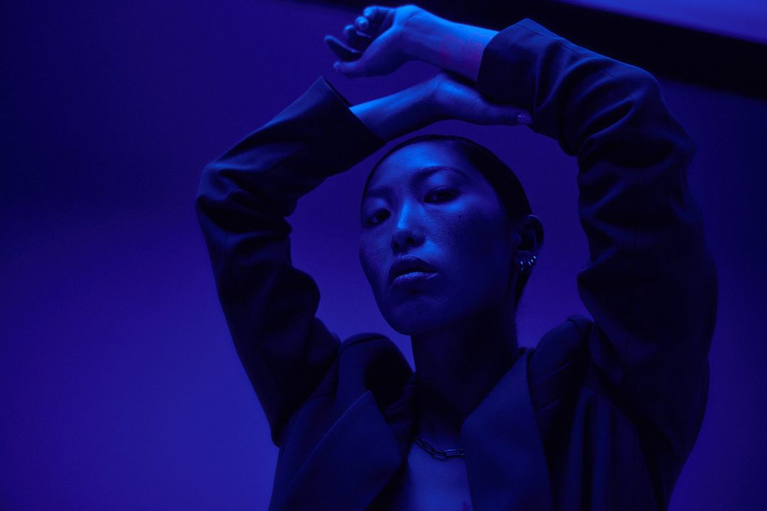 Portrait of an asian adult woman in a black blazer with her hands above her head and illuminated by blue lighting in a studio.