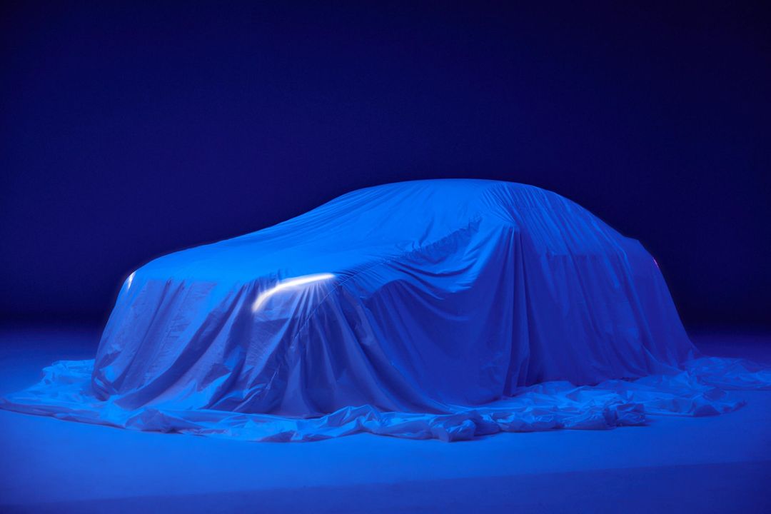Blue-toned image of a car draped in cloth, revealing headlights, suggesting a new model debut in the automotive industry.