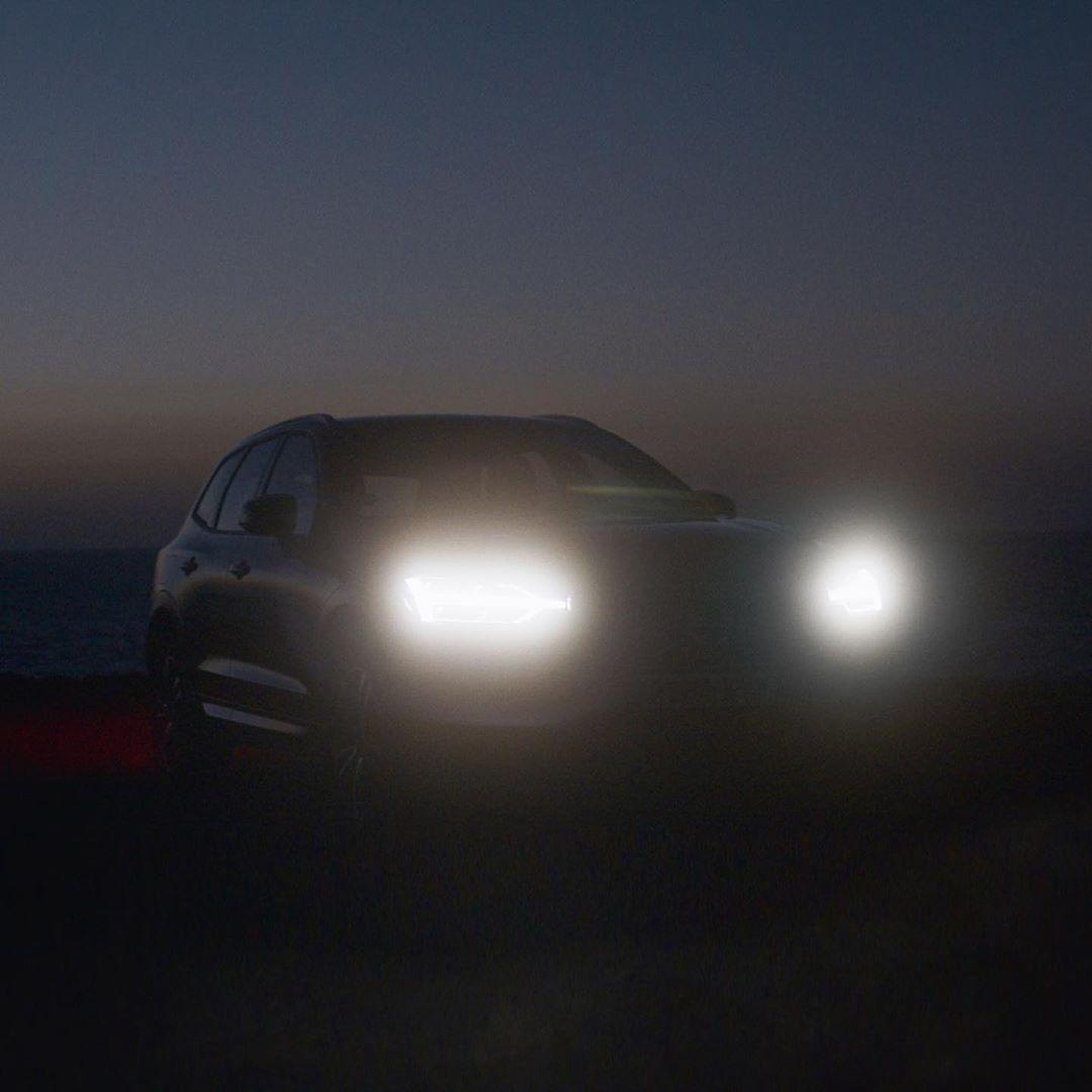 Dark automotive shot of a gray SUV with illuminated headlights at dusk near the ocean, creating a sense of mystery and adventure.