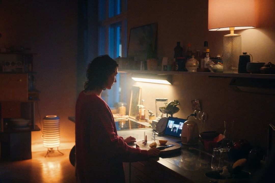Woman prepares food in a warmly lit kitchen. Video call screen visible with people. Interior shows a home kitchen with soft lighting.