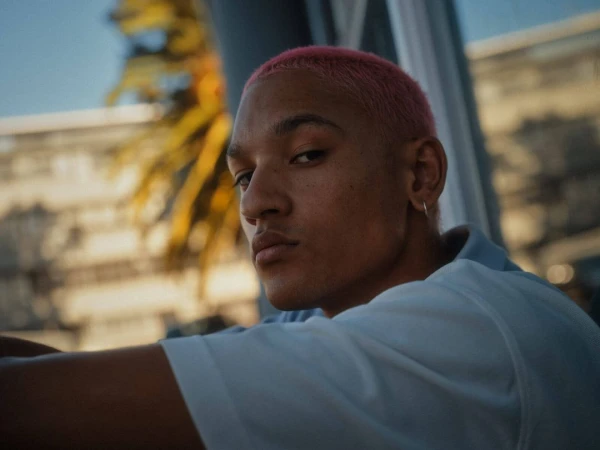 Close-up portrait of a young man with pink short-cropped hair wearing a light blue polo shirt, looking over his shoulder.
