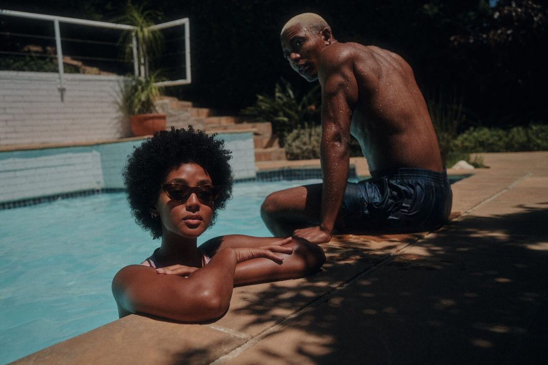 Woman with afro in swimming pool near man. Dark-skinned adult duo relaxing by the pool on a summer day with greenery background.