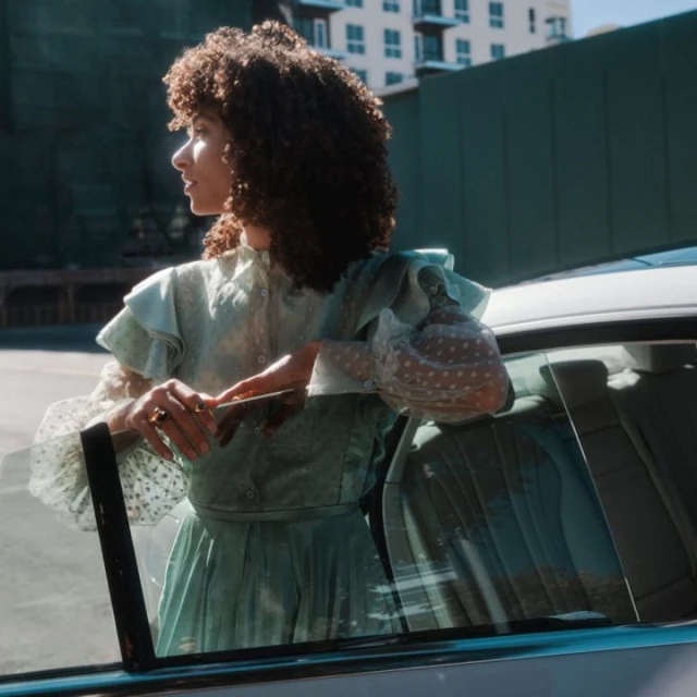 A curly-haired woman in a vintage-style pale green dress is leaning against the open door of a car outdoors on a sunny day.