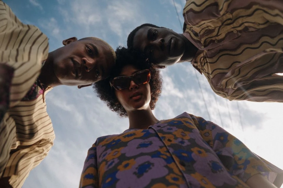 Three young adults in patterned tops look downward, the central figure wearing sunglasses. Blue sky and wisps of clouds fill the background.