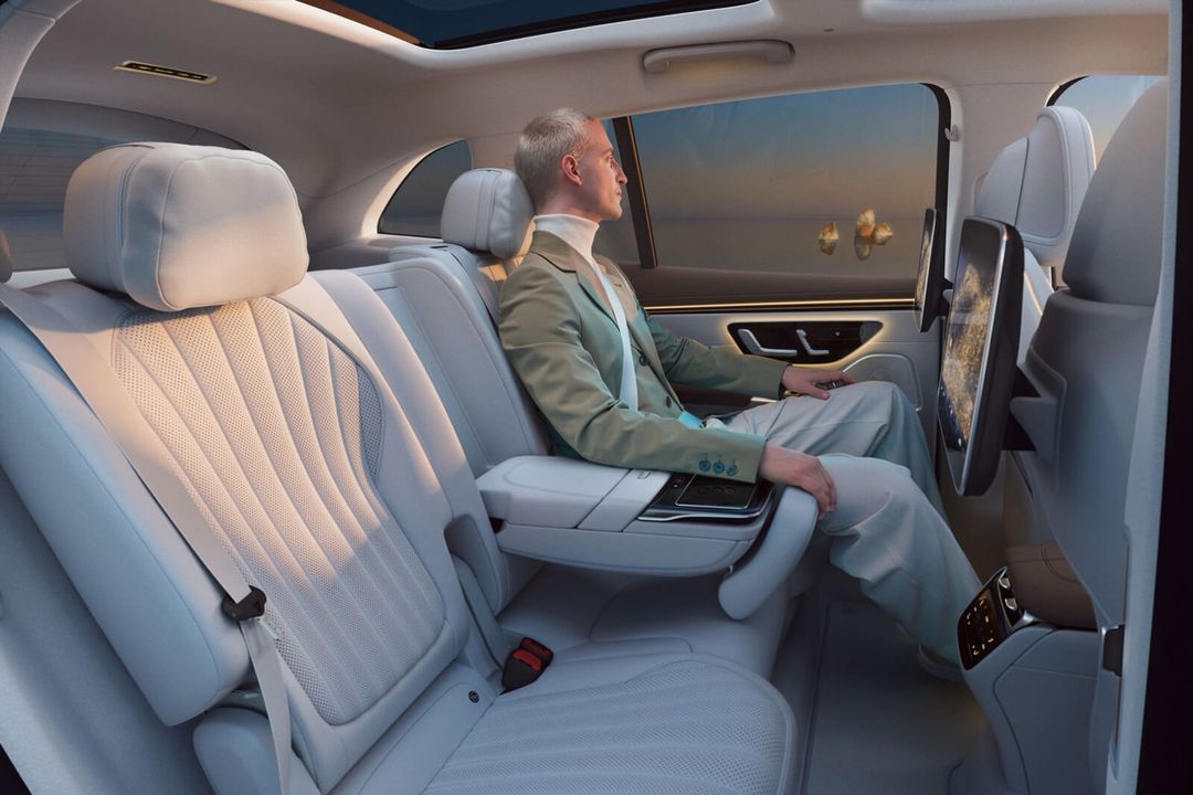Man in a blazer sits in the back of a luxury car with white leather seats, looking out the window at a landscape with trees at sunset.