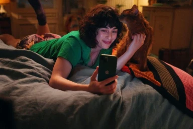 Woman with curly dark hair lying on a bed in a green t-shirt holding a smartphone while an orange cat nuzzles her face.