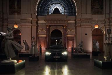 A dark electric car parked inside a grand hall featuring arched doorways, ornate columns, and classical statues on display pedestals.