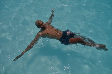 Man with short blonde-dyed hair floating on his back in clear blue water wearing dark blue swim trunks