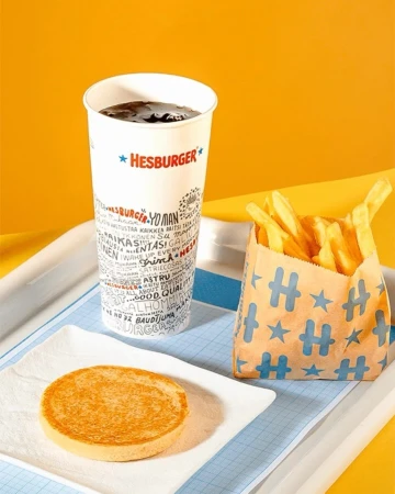 Hesburger meal on a tray: soda cup with logo, fries in a branded bag, and burger bun on a small square white plate against yellow.