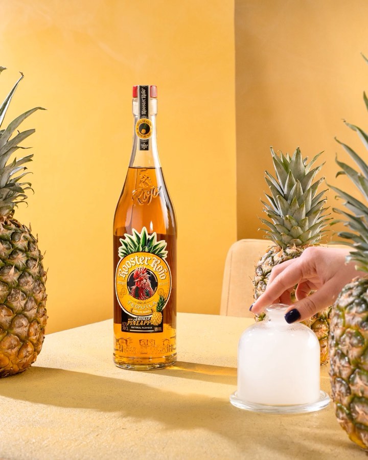 Rooster Rojo pineapple flavored tequila bottle surrounded by pineapples and a cloche filled with smoke in a studio setting.