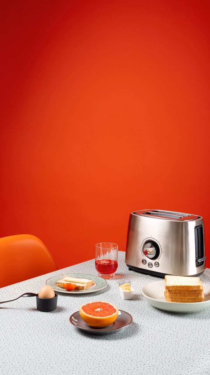 Still life shot of a chrome toaster on a table with toast, grapefruit, and a glass of juice against a red studio background.