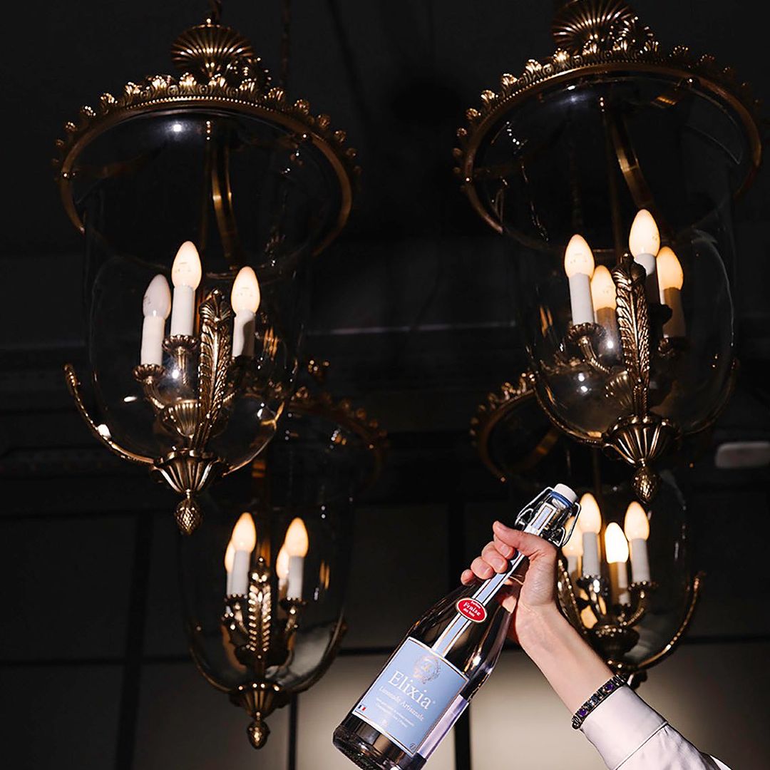 Hand holds a bottle of Elixia soda, set against chandeliers with lit bulbs. The scene has a dark, elegant tone and ambience.