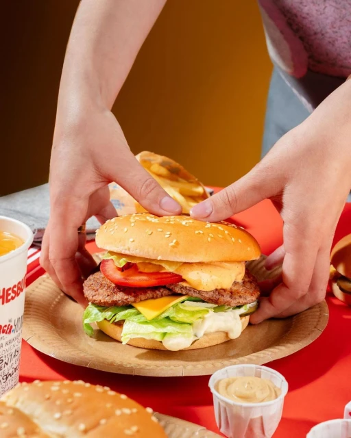 Hands assembling a tall burger with layers of lettuce, tomato, cheese, and a sesame seed bun on a paper plate, set on a red surface.