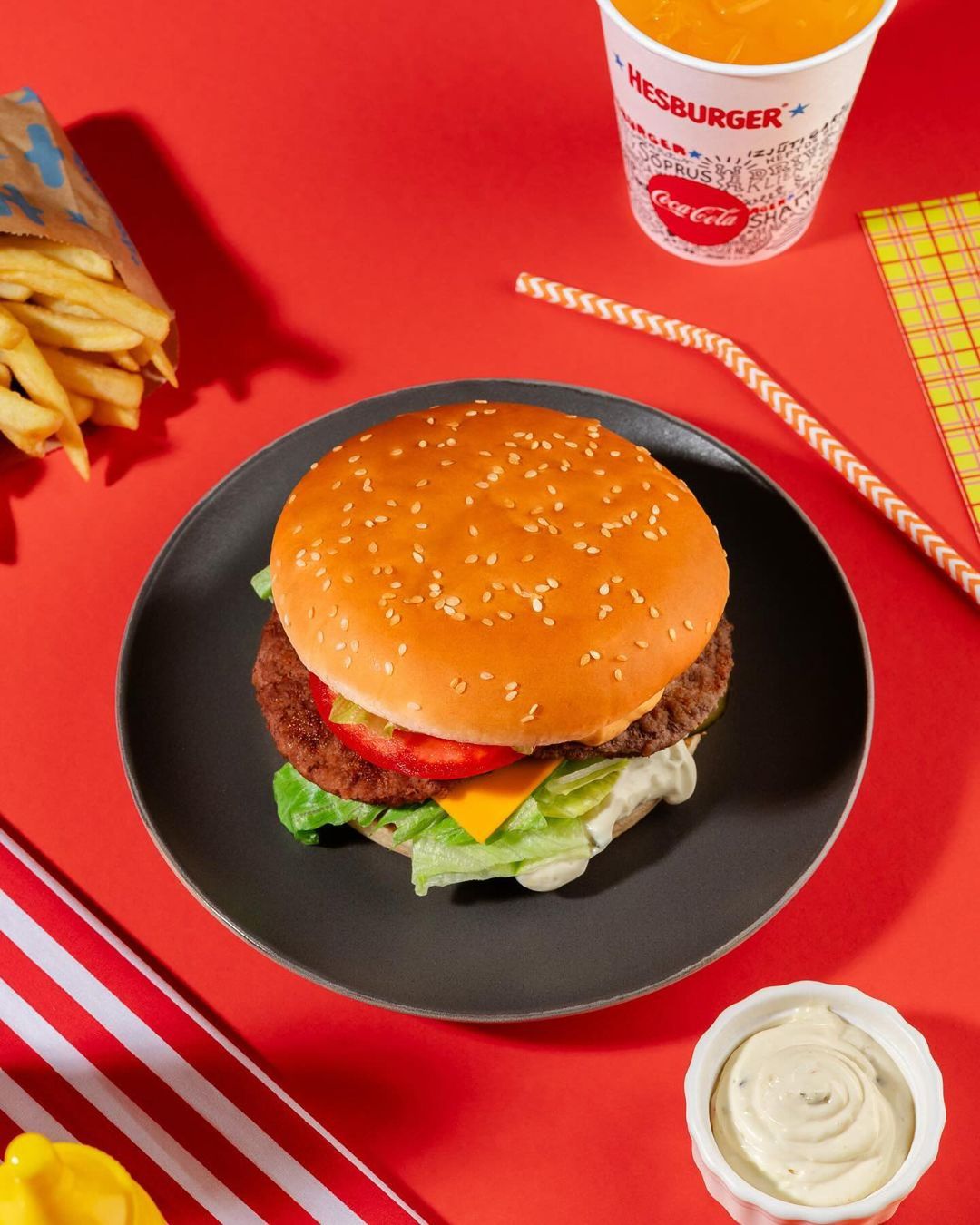 A burger on a black plate sits on a red surface, accompanied by fries, a drink, sauce, and a striped cloth. Food photography.