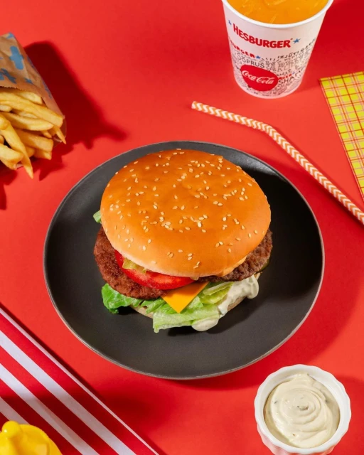 A burger on a black plate sits on a red surface, accompanied by fries, a drink, sauce, and a striped cloth. Food photography.