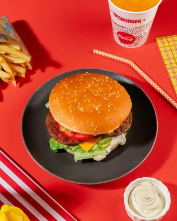 A burger on a black plate sits on a red surface, accompanied by fries, a drink, sauce, and a striped cloth. Food photography.