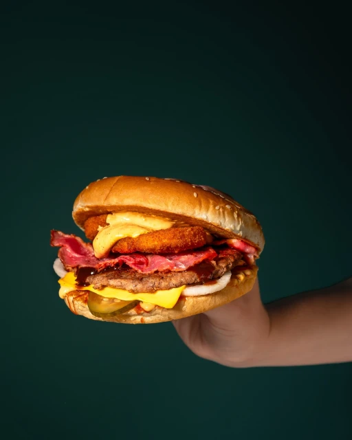 Hand holds a large, stacked hamburger featuring layers of meat, cheese, bacon, and onion rings against a dark green background.