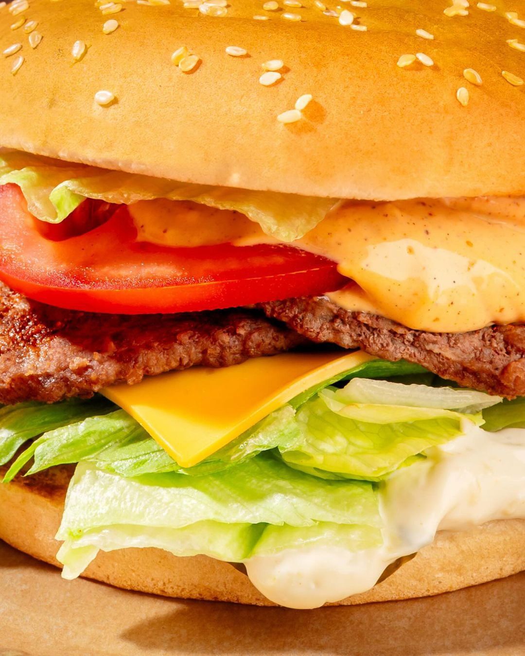 Close-up of a burger featuring a sesame seed bun, beef patty, cheese, tomato, lettuce, and a creamy sauce, showcasing the food's textures.