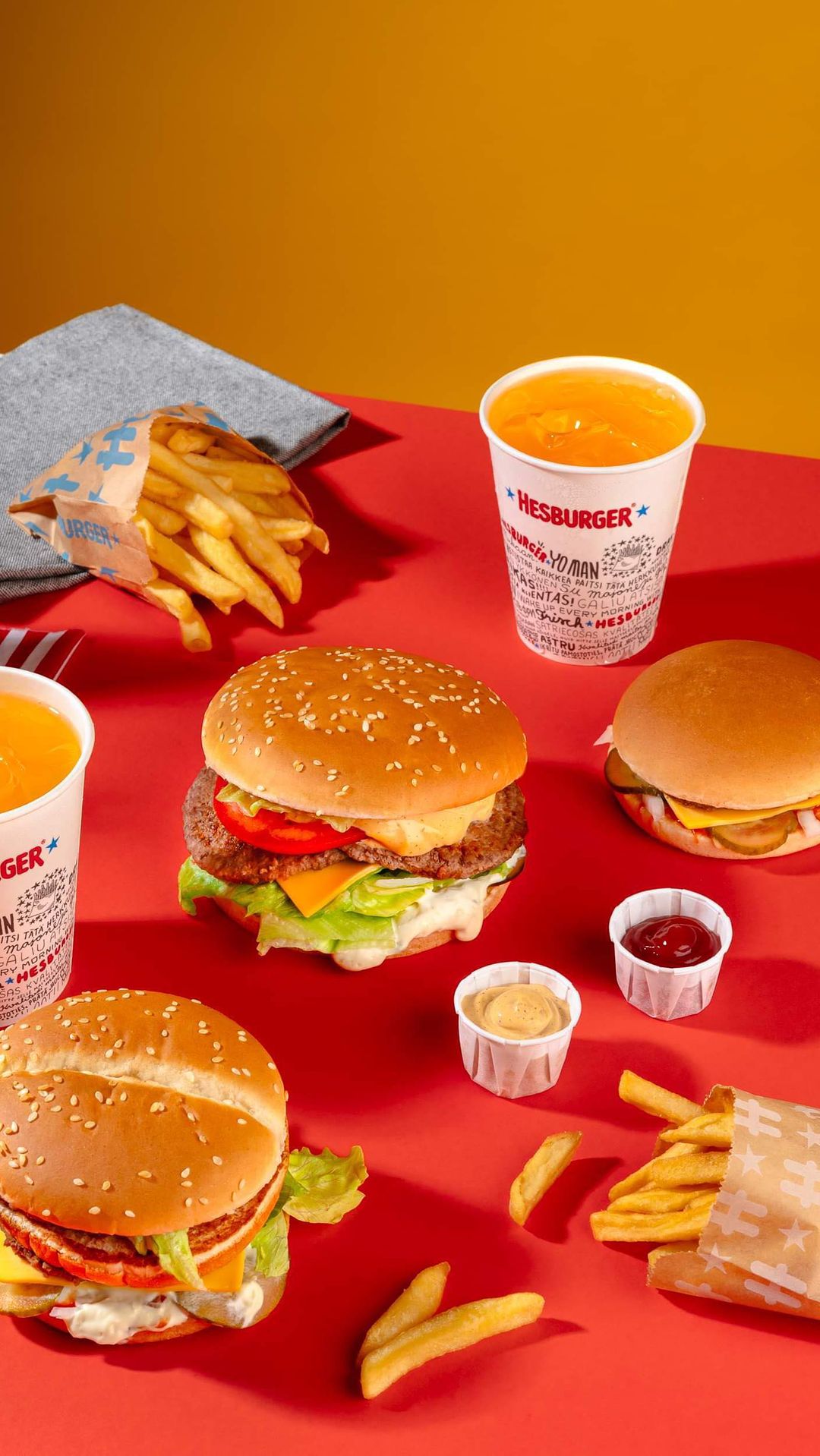 Overhead shot of burgers, fries, and orange drinks arranged on a red surface with a mustard-colored backdrop; food and beverage.