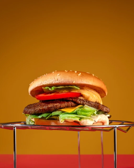 Studio shot of a double cheeseburger with lettuce, tomato, cheese, and sauce on a metal rack, set against a gradient orange background.