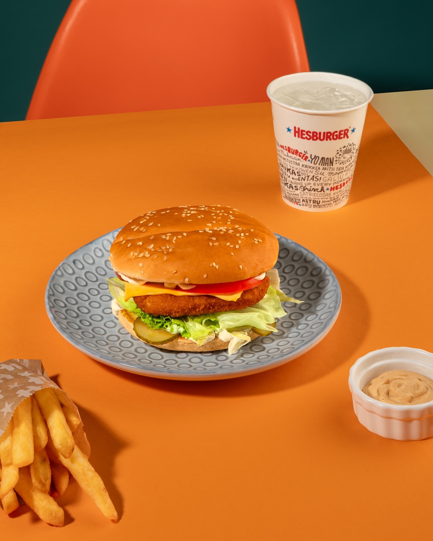 A sesame seed bun burger with tomato, lettuce, and pickles sits on a plate with fries, dip, and a drink, set against an orange backdrop.