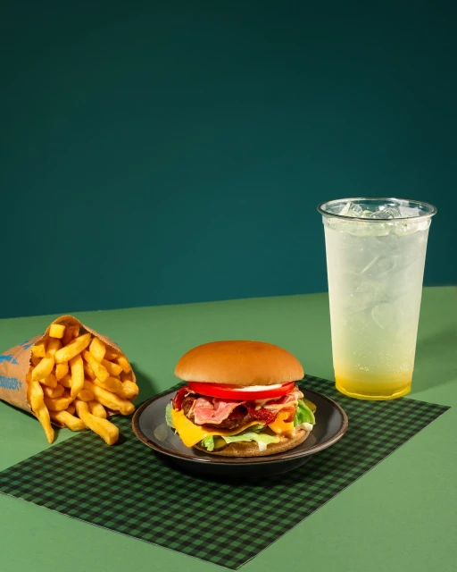 Overhead shot of a burger, french fries, and a lemon drink on a checkered surface with a green background.