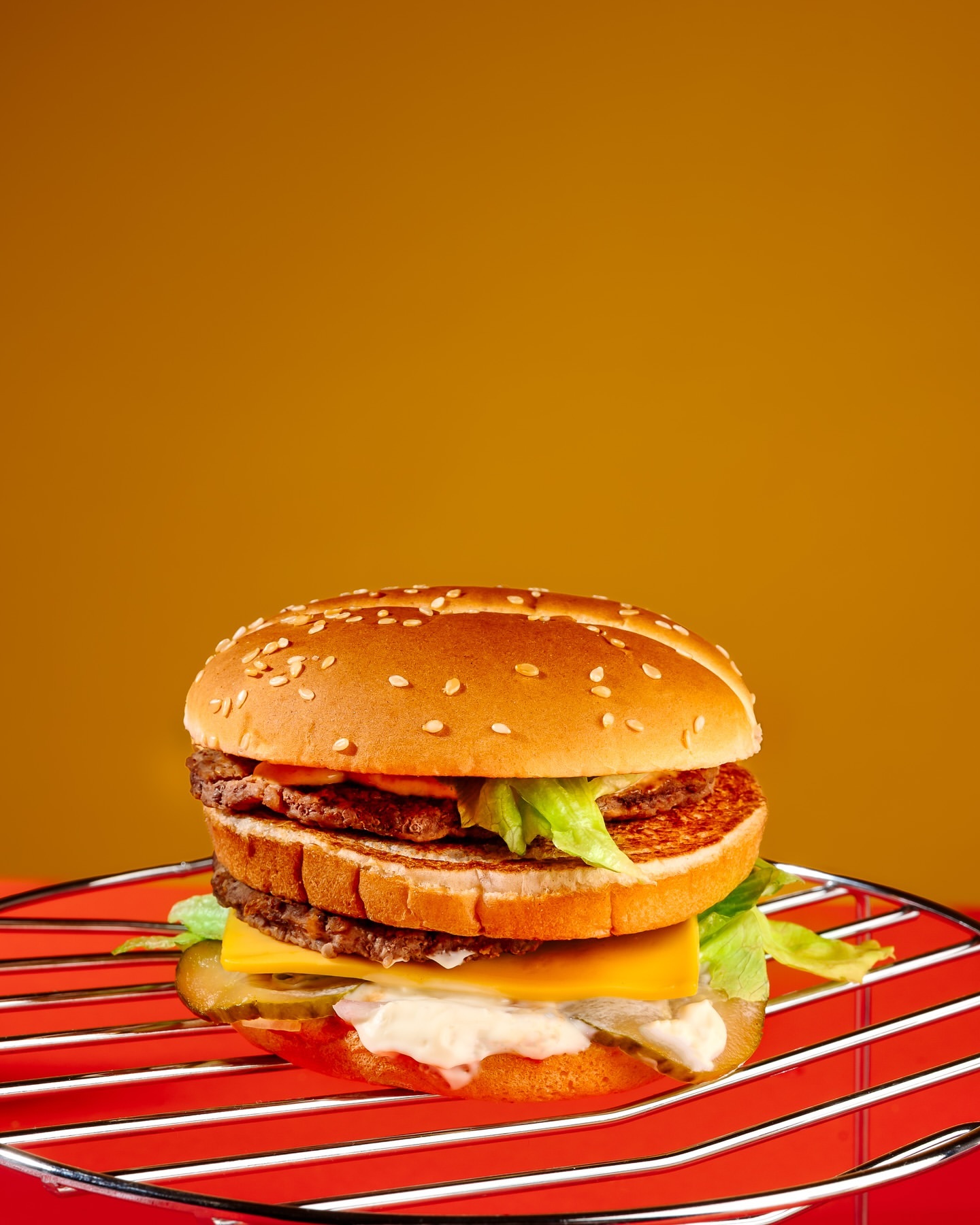 Two-tiered hamburger with beef patties, cheese, lettuce, and sauce on a sesame bun, presented on a red grill rack against a warm gradient background.