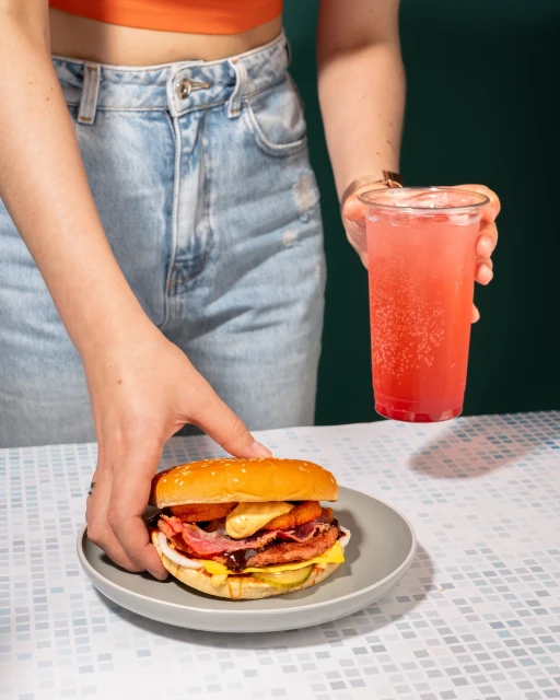 A person is holding a burger on a plate and a drink, with a green background. The scene is well-lit, highlighting the burger's details.