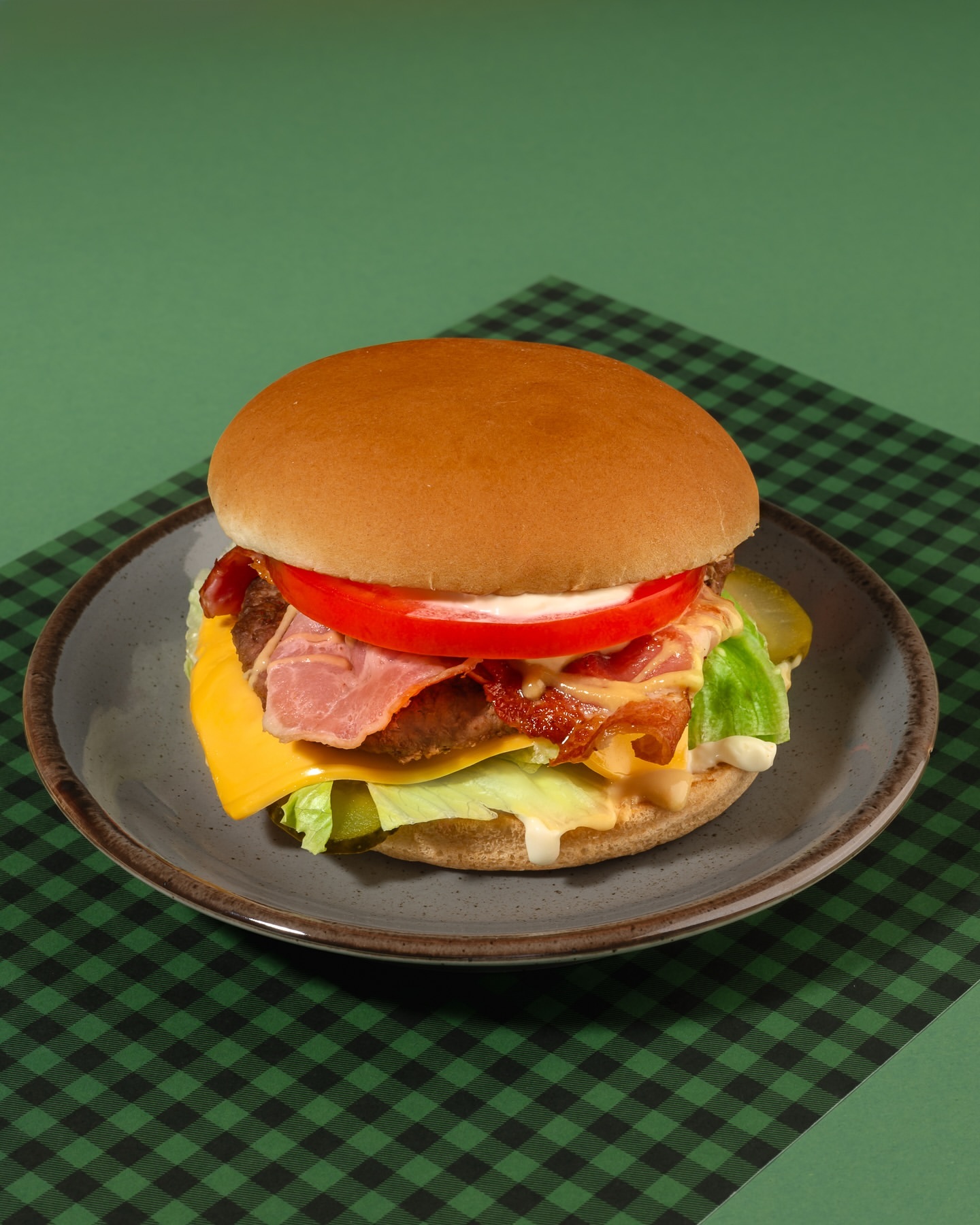 Close-up of a deli-style burger with bacon, cheese, tomato, lettuce, and pickles on a grey plate, set against a green background.