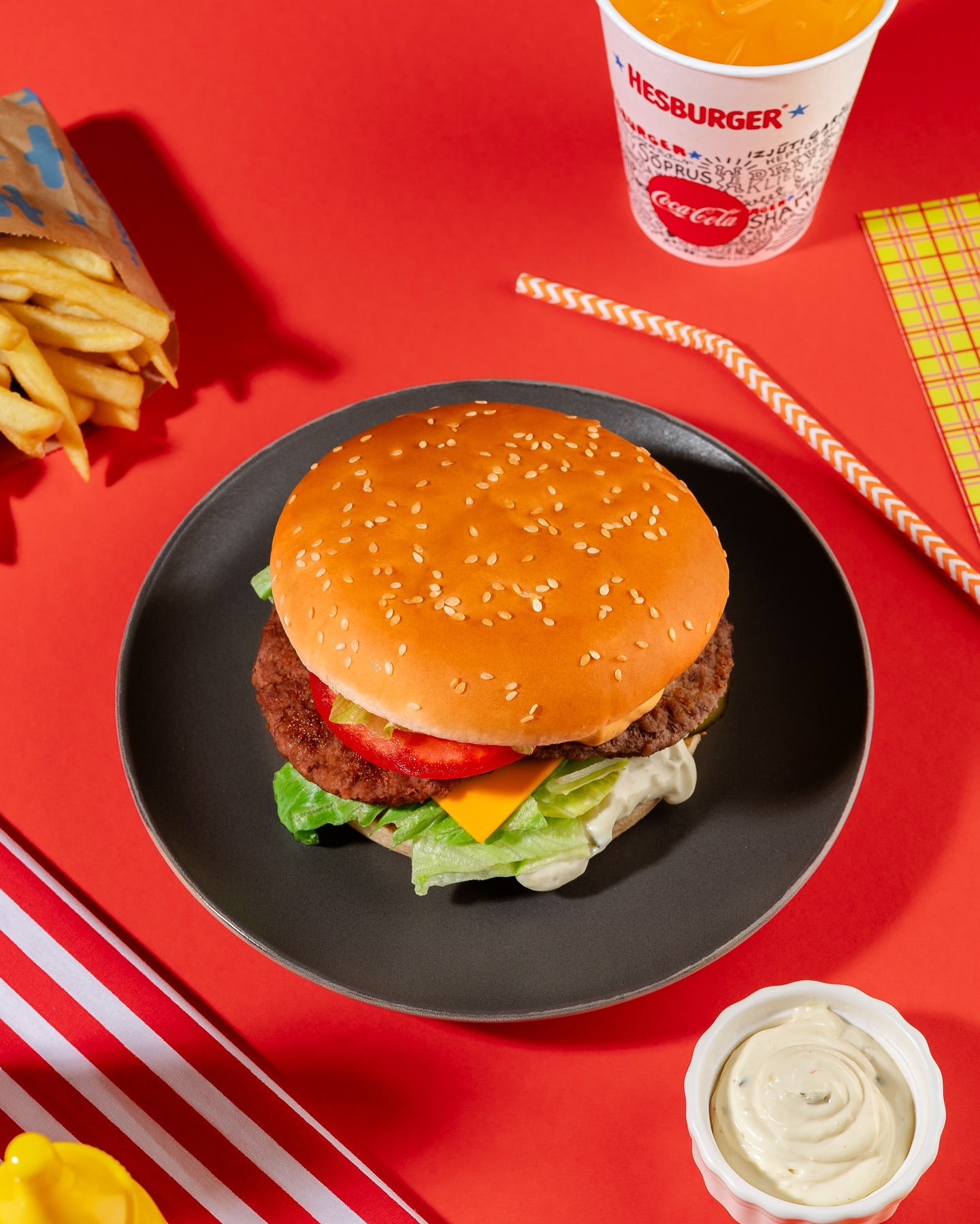 Overhead shot of a Hesburger hamburger on a plate with fries, drink, sauce, and red background, commercial food photography.
