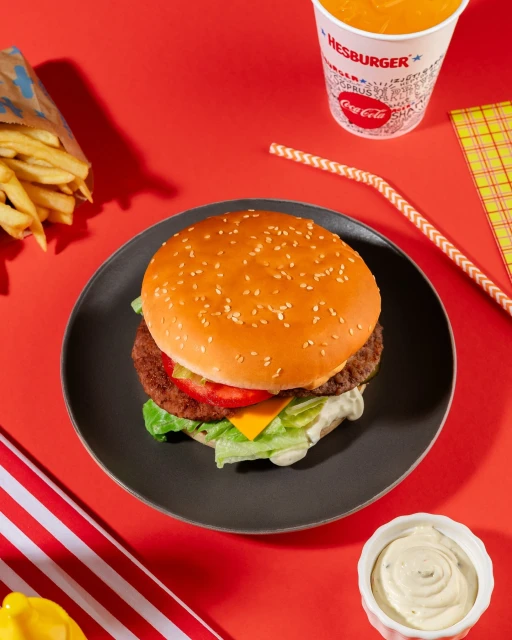 Overhead shot of a Hesburger hamburger on a plate with fries, drink, sauce, and red background, commercial food photography.