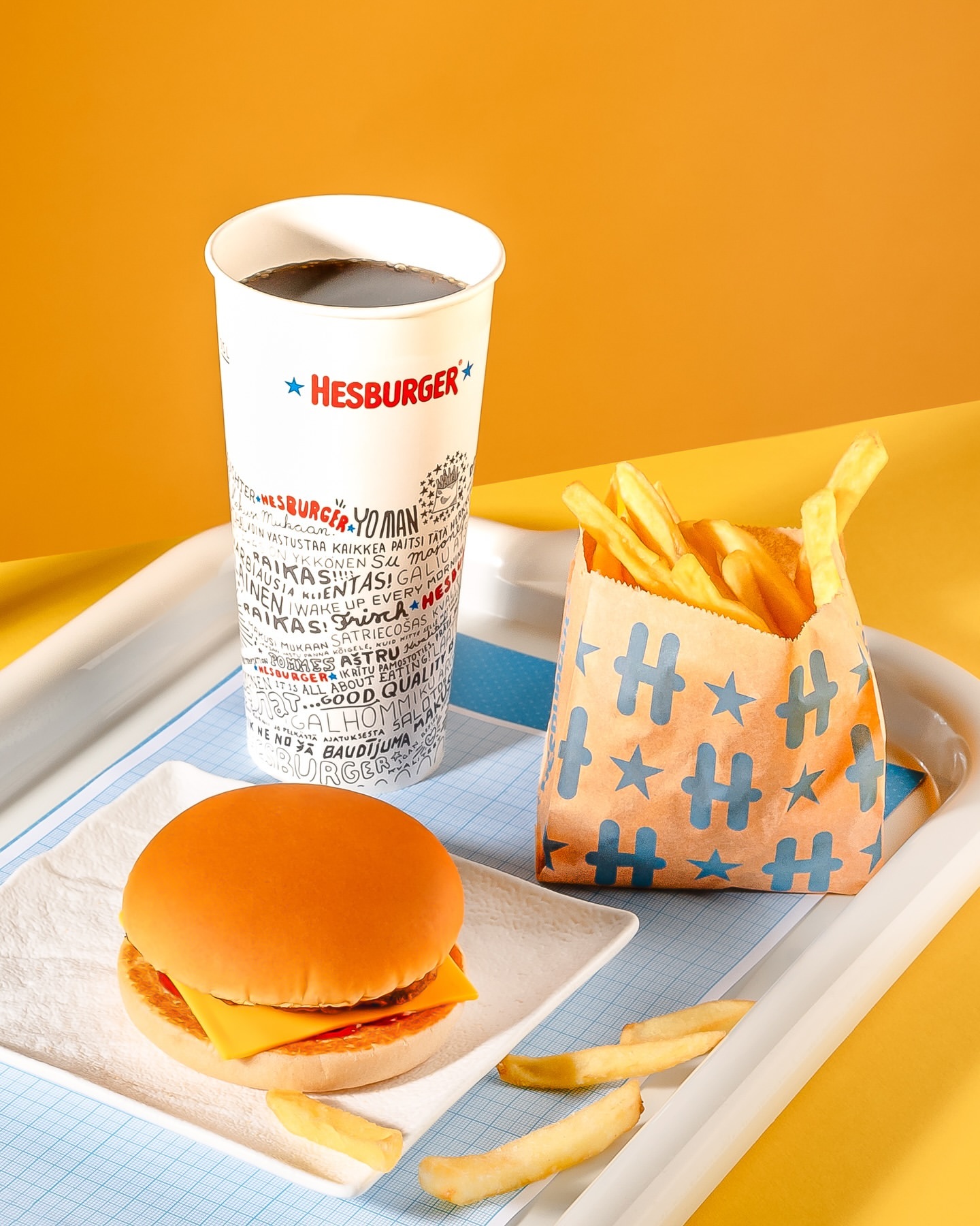 Still life shot of a cheeseburger on a white plate with a bag of fries and a soda on a tray against a yellow and orange background.