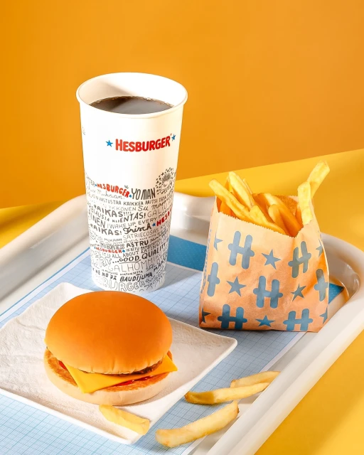 Still life shot of a cheeseburger on a white plate with a bag of fries and a soda on a tray against a yellow and orange background.