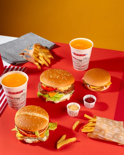 Overhead shot of a vibrant fast food spread with burgers, fries, and orange drinks against a split red and mustard-yellow backdrop.