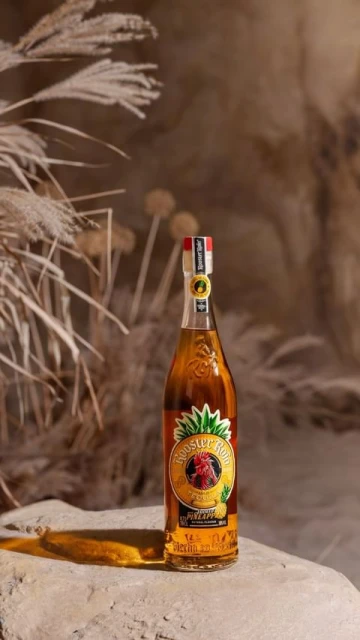 Bottle of Rooster Rojo pineapple flavored tequila on a rock surface, with reeds and tan backdrop, product photography.