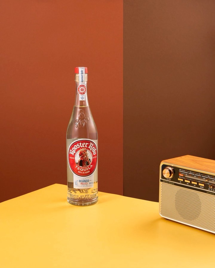 A bottle of Rooster Rojo Tequila on a yellow table paired with a wooden radio against a split orange and brown backdrop.