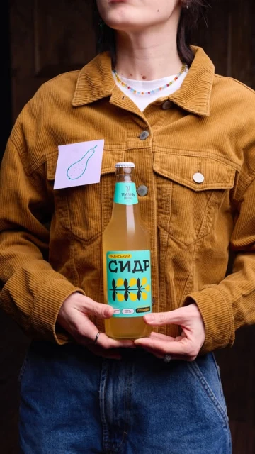 Young adult presents a bottle of pear cider in front of a brown wooden background, adorned with a pear drawing and wearing a brown jacket.